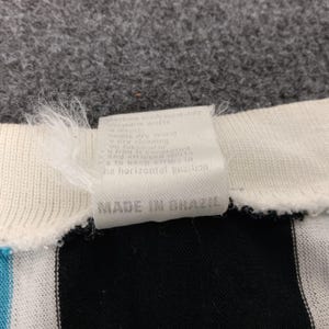 May include: A white tag with black text that reads "Made in Brazil" attached to a black and white striped shirt. The tag also includes washing instructions in a smaller font.