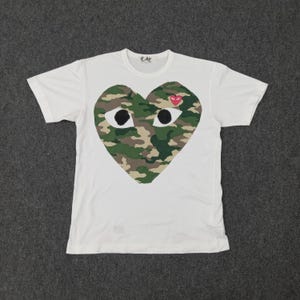 Puede incluir: Camiseta blanca con un gran diseño de camuflaje en forma de corazón. El corazón tiene dos formas de ojos negros y un pequeño corazón rojo. La palabra "PLAY" está impresa en el cuello.