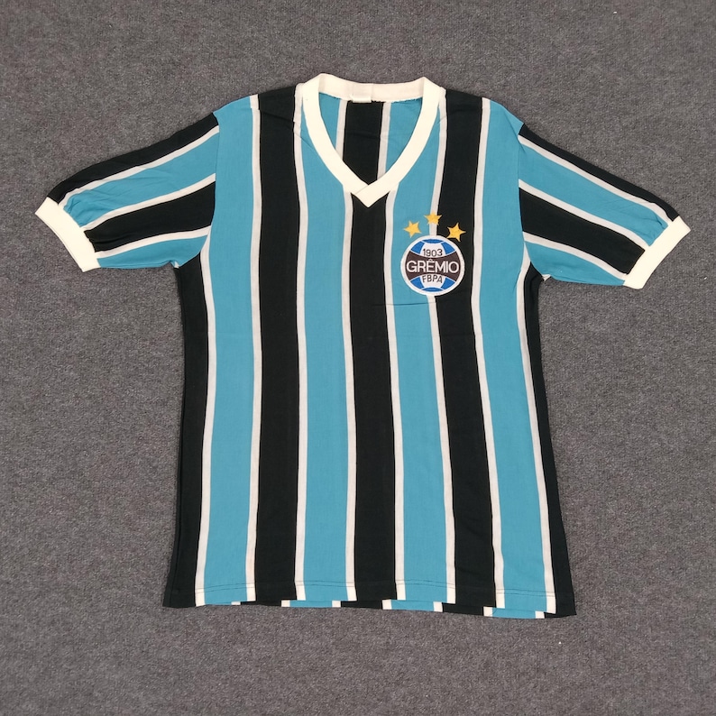 May include: A blue and black striped soccer jersey with a white v-neck collar. The jersey has a circular patch with the text "1903 GREMIO FBPA" and three gold stars.
