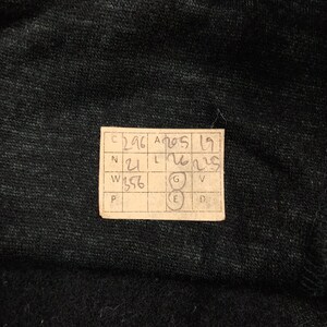 May include: A dark charcoal gray fabric with a textured, heathered appearance. A rectangular paper tag is attached, featuring a grid with handwritten numbers and letters including C 296, A 205, 19, N 21, L 26, 225, W 356, G, E, V, P, and D.