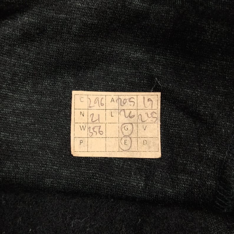 May include: A dark charcoal gray fabric with a textured, heathered appearance. A rectangular paper tag is attached, featuring a grid with handwritten numbers and letters including C 296, A 205, 19, N 21, L 26, 225, W 356, G, E, V, P, and D.