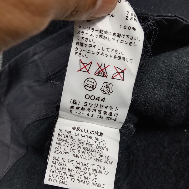 May include: A white fabric garment care label attached to a dark gray textile. The label features black printed text in Japanese, French, and English, including care instructions, symbols, and the manufacturer name Yohji Yamamoto Inc.