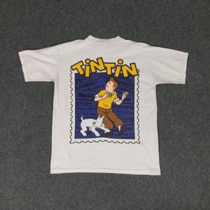 May include: White t-shirt featuring a graphic of Tintin, a cartoon character, and his dog Snowy. The design is set within a blue postage stamp frame, with the word "TINTIN" in yellow at the top. The shirt is made of a soft, comfortable material.