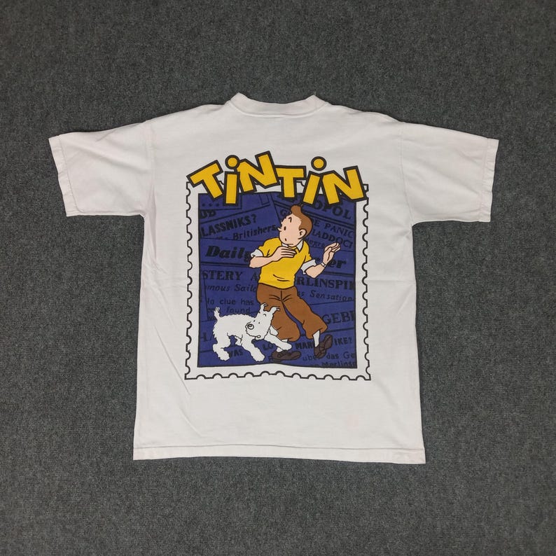 May include: White t-shirt featuring a graphic of Tintin, a cartoon character, and his dog Snowy. The design is set within a blue postage stamp frame, with the word "TINTIN" in yellow at the top. The shirt is made of a soft, comfortable material.
