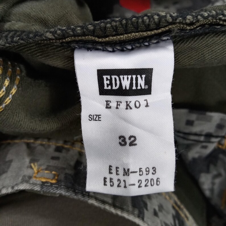 Op de afbeelding: Een wit label met zwarte tekst dat "EDWIN. EFKO1 SIZE 32 EEM-593 E521-2206" aangeeft.