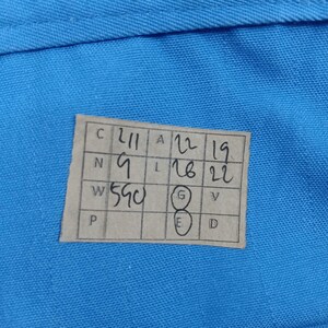 Puede incluir: Primer plano de una tela azul claro con una etiqueta rectangular. La etiqueta tiene n&uacute;meros y letras manuscritos, incluyendo "211", "9" y "590". N&uacute;meros adicionales est&aacute;n dispuestos en columnas.