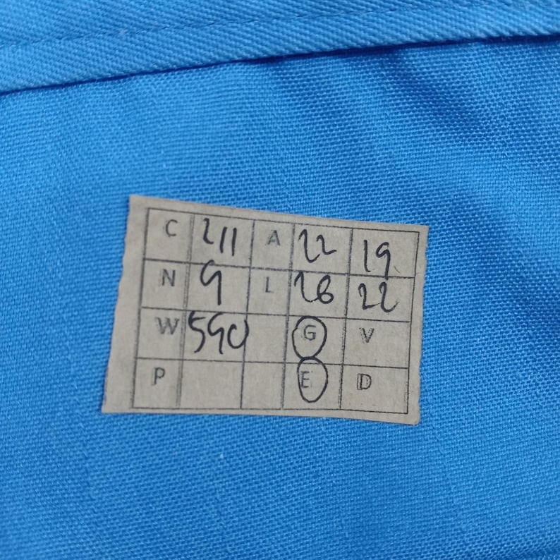 Puede incluir: Primer plano de una tela azul claro con una etiqueta rectangular. La etiqueta tiene n&uacute;meros y letras manuscritos, incluyendo "211", "9" y "590". N&uacute;meros adicionales est&aacute;n dispuestos en columnas.