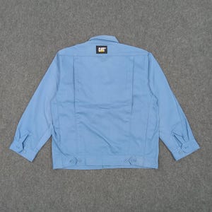 Puede incluir: Chaqueta de trabajo azul claro con botones y cuello. La parte trasera de la chaqueta presenta un peque&ntilde;o parche con el logotipo "CAT" en amarillo y negro. La chaqueta tiene mangas largas con pu&ntilde;os abotonados.