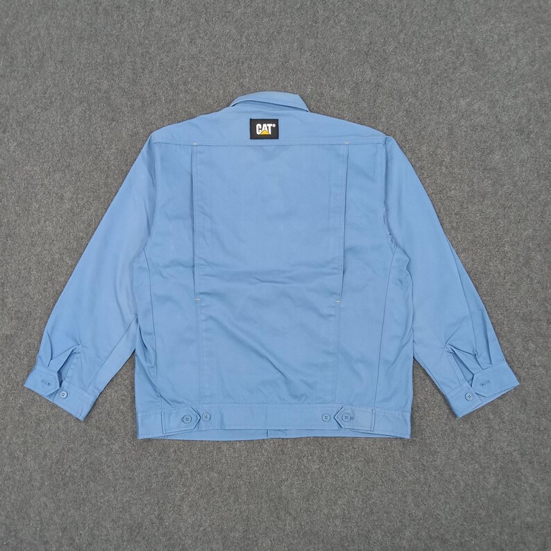Puede incluir: Chaqueta de trabajo azul claro con botones y cuello. La parte trasera de la chaqueta presenta un peque&ntilde;o parche con el logotipo "CAT" en amarillo y negro. La chaqueta tiene mangas largas con pu&ntilde;os abotonados.