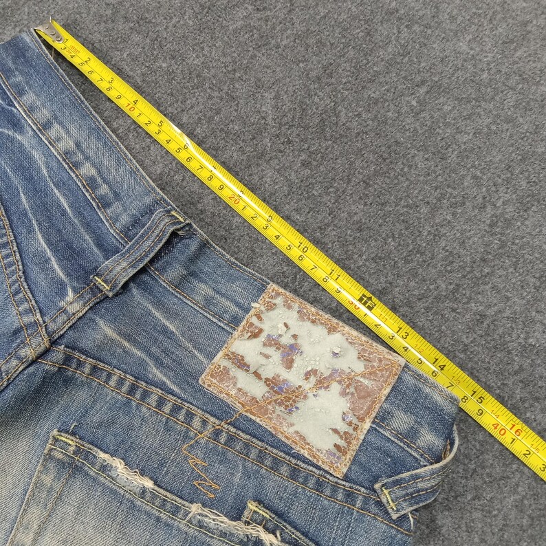 Op de afbeelding: Close-up van een blauwe denim jeans met een distressed look. Een gele meetlint ligt over de jeans, met metingen in centimeters. De jeans heeft een bruine leren patch op de achterzak.