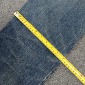 以下が含まれることがあります： 青いデニム生地のクローズアップと黄色の巻尺。テープはセンチメートル単位の測定値を示し、デニムの質感と色を強調しています。巻尺は生地の上で角度がついています。