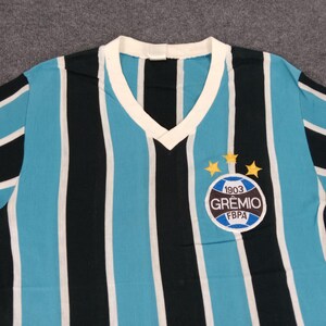May include: A blue and black striped soccer jersey with a white v-neck collar. The jersey has a circular patch with the text "1903 GR&Ecirc;MIO FBPA" and two gold stars above it.