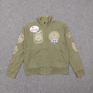 May include: Olive green zip-up jacket with multiple embroidered patches. The jacket features a high collar, two front pockets, and the "Avirex Corp" logo patch. Patches include military-themed designs and text.