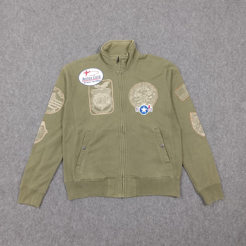 May include: Olive green zip-up jacket with multiple embroidered patches. The jacket features a high collar, two front pockets, and the "Avirex Corp" logo patch. Patches include military-themed designs and text.