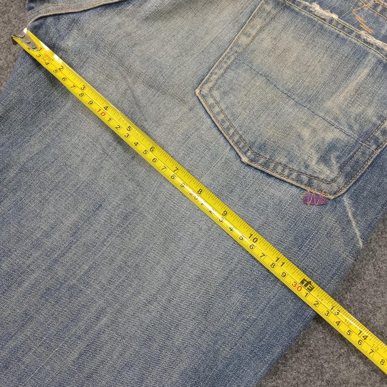 Op de afbeelding: Close-up van lichtblauwe denim jeans met een achterzak. Een gele meetlint ligt over de jeans en toont metingen in centimeters. De jeans heeft een versleten uiterlijk met wat beschadigingen.