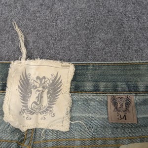 May include: Close-up of denim jeans with a light blue wash. The jeans feature two tags with a winged emblem and the letter "J". One tag is square and frayed, the other is rectangular with the size "34".