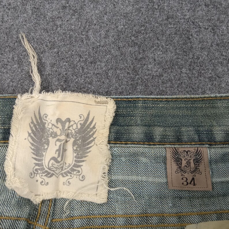 May include: Close-up of denim jeans with a light blue wash. The jeans feature two tags with a winged emblem and the letter "J". One tag is square and frayed, the other is rectangular with the size "34".