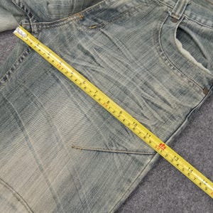 May include: Close-up of light blue denim jeans with a yellow measuring tape showing measurements in inches. The jeans have a faded wash and visible pocket details. The measuring tape shows measurements up to 14 inches.