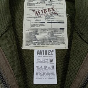 May include: Close-up of an olive green zip-up hoodie with a tag featuring the brand "AVIREX" and care instructions. The tag also includes the text "100% COTONE BAUMWOLLE COTON-COTTON". The hoodie is made of cotton.