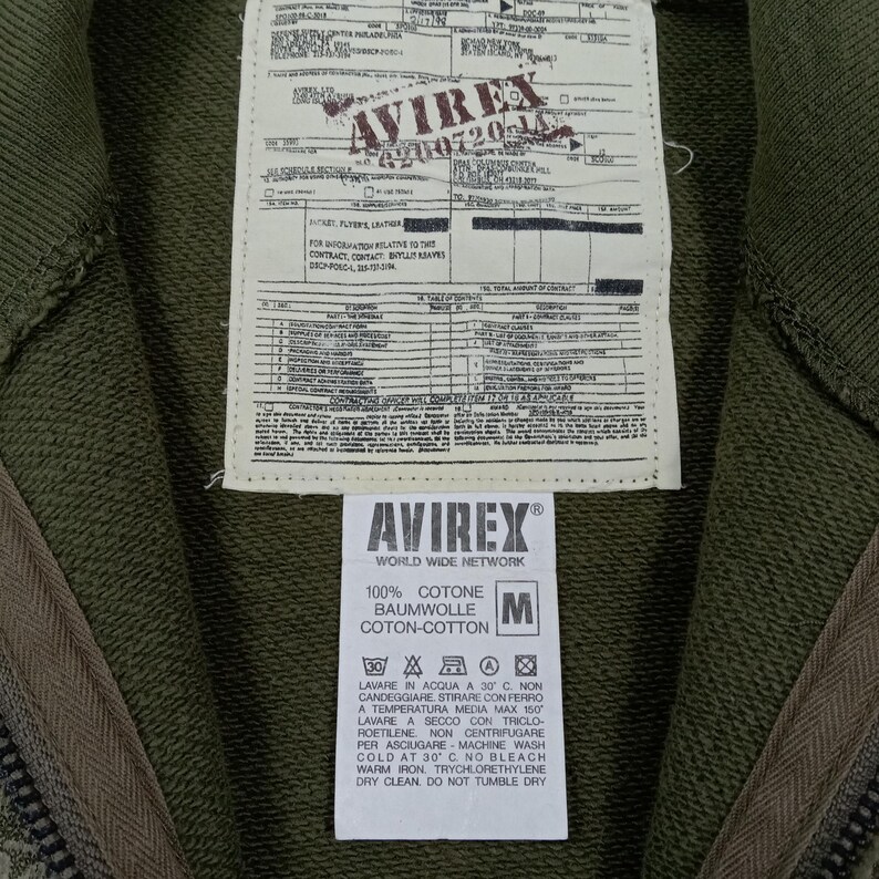 May include: Close-up of an olive green zip-up hoodie with a tag featuring the brand "AVIREX" and care instructions. The tag also includes the text "100% COTONE BAUMWOLLE COTON-COTTON". The hoodie is made of cotton.