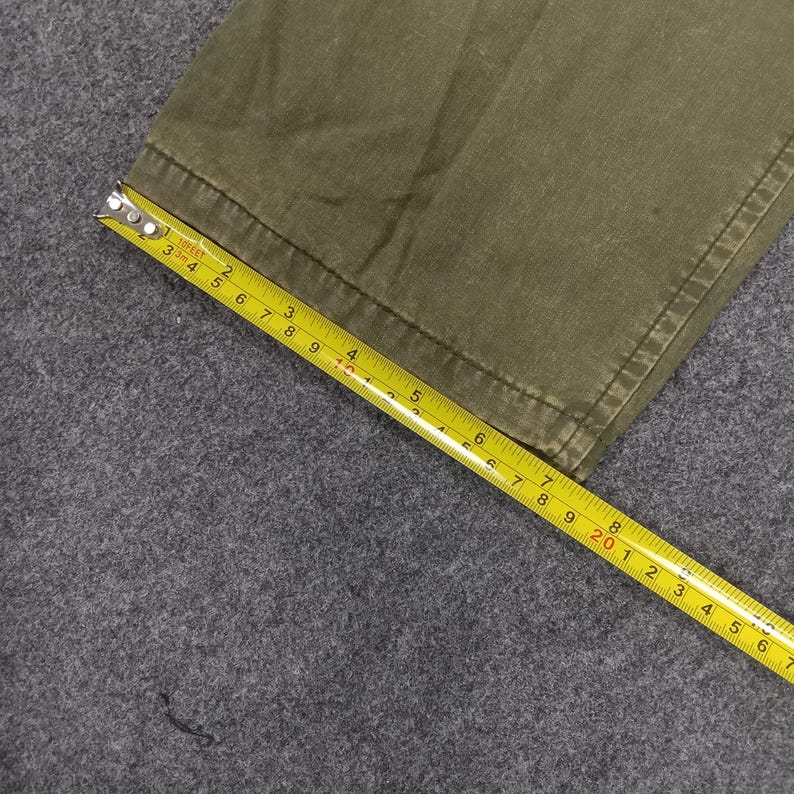 May include: Close-up of the bottom edge of a pair of olive green cargo trousers. A yellow measuring tape is laid across the fabric.