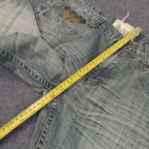 May include: Light blue denim jeans with a faded wash and distressed details. A yellow measuring tape is laid across the jeans, showing measurements in inches. The jeans have a decorative pocket design.