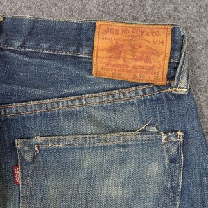 以下が含まれることがあります： バックポケットと茶色の革パッチが付いた、青いデニムジーンズのクローズアップ。パッチには「JOE McCOY & CO.」と書かれており、衣類の保証に関するテキストが含まれています。ジーンズのサイズはW29 L36、ウエスト約74cm、丈約91cmです。