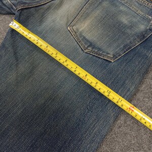 以下が含まれることがあります： 黄色いメジャーで測定された青いデニムジーンズのクローズアップ。テープはセンチメートル単位の測定値を示し、生地の質感と衣服の詳細を強調しています。ジーンズにはバックポケットが付いています。