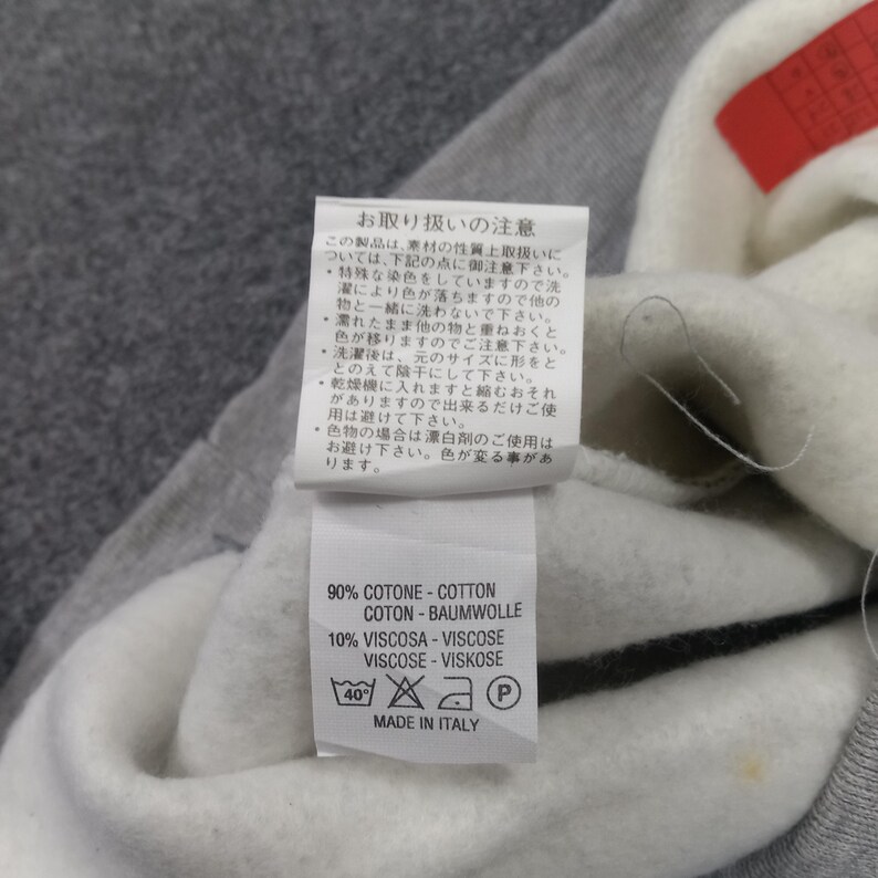 May include: Close-up of a white tag with black text and care symbols, attached to a gray and white garment. The tag reads "90% COTONE-COTTON, COTON-BAUMWOLLE, 10% VISCOSA-VISCOSE, VISCOSE-VISKOSE, MADE IN ITALY."