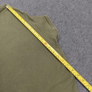 May include: Olive green fleece jacket with a small square pocket and a yellow measuring tape laid across it. The tape shows measurements in inches, indicating the jacket's dimensions. The jacket is made of a soft, textured material.