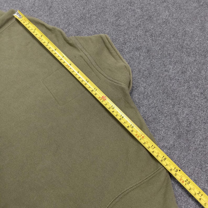 May include: Olive green fleece jacket with a small square pocket and a yellow measuring tape laid across it. The tape shows measurements in inches, indicating the jacket's dimensions. The jacket is made of a soft, textured material.