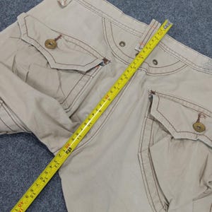 May include: Beige cargo pants with multiple pockets and brown stitching. A measuring tape is laid across the pants.