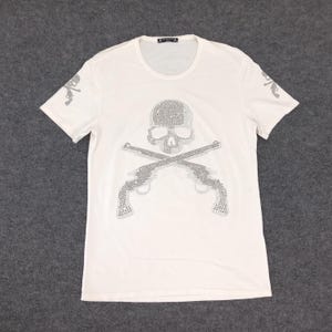 May include: White t-shirt with a skull and crossbones design made of rhinestones. The skull is wearing a bandana.