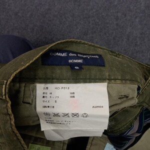 May include: Green cargo trousers with a Comme des Gar&ccedil;ons Homme label. The label features the brand name, Homme, and the size S. The care label includes the brand name, the style number HO-P013, and the fabric content: 100% cotton and 100% cupro. The care instructions are in Japanese.