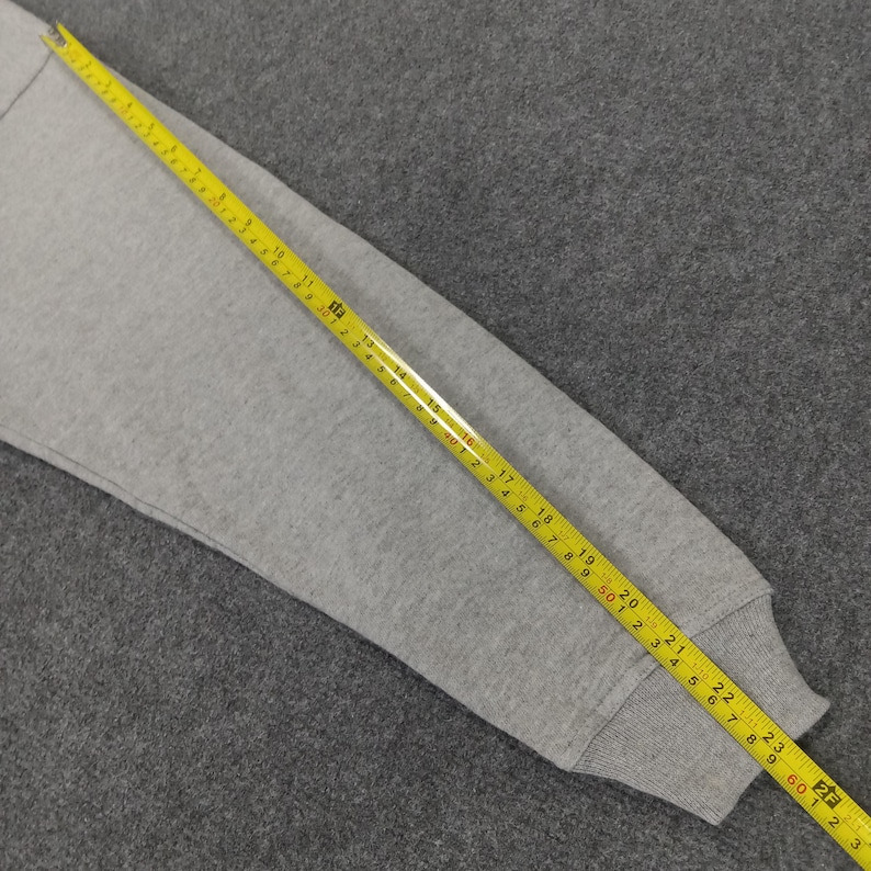 May include: A gray sweatshirt sleeve with a yellow measuring tape laid across it. The tape shows measurements in inches, with the sleeve's cuff at the 23-inch mark. The fabric is a solid, light gray color.