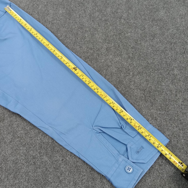 Puede incluir: Camisa azul claro de manga larga con pu&ntilde;o abotonado y tapeta abotonada. Una cinta m&eacute;trica amarilla se coloca sobre la camisa, mostrando medidas en cm.