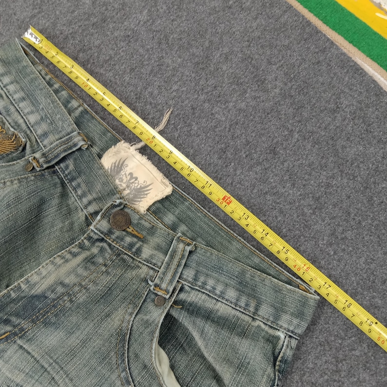 May include: Light blue denim jeans with a button closure and a decorative label. A yellow measuring tape is laid across the jeans, showing measurements in inches. The jeans are on a gray surface.