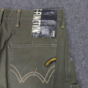 Op de afbeelding: Groene cargo broek met een bruin leren patch waarop "E-FUNCTION" en "EDWIN" in witte tekst staat. De broek heeft een geel en bruin stikdetail op de achterzakken.
