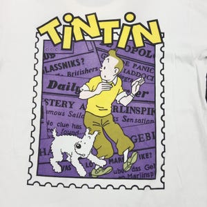 May include: White t-shirt featuring a graphic of Tintin and Snowy from the Tintin comic series. The design includes the word "TINTIN" in yellow, a purple background with newspaper clippings, and a stamp-like border.