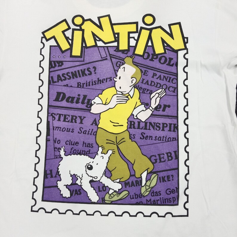May include: White t-shirt featuring a graphic of Tintin and Snowy from the Tintin comic series. The design includes the word "TINTIN" in yellow, a purple background with newspaper clippings, and a stamp-like border.