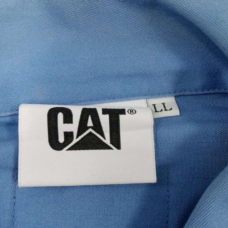 Puede incluir: Camisa de trabajo azul claro con el logotipo de CAT en blanco y negro sobre una etiqueta blanca. La camisa tiene cuello abotonado y una etiqueta que dice "LL".