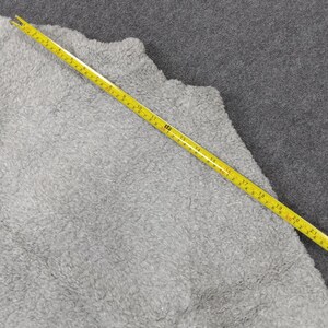 Op de afbeelding: Een close-up van een grijze, pluche stof met een gele meetlint erop. De tape meet 50 cm.