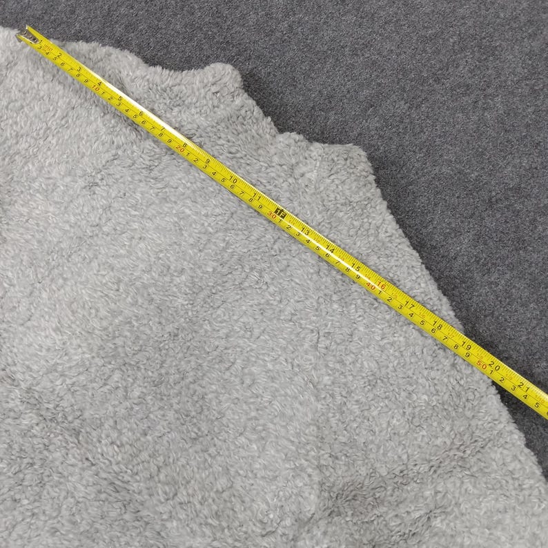 Op de afbeelding: Een close-up van een grijze, pluche stof met een gele meetlint erop. De tape meet 50 cm.
