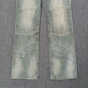 May include: Faded light blue denim jeans with a patchwork design on the knees and lower legs. The jeans have a distressed look with visible wear and tear, giving them a vintage aesthetic. The jeans are laid flat on a gray surface.