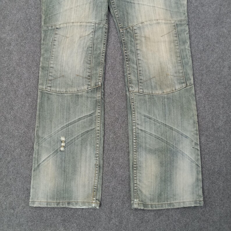 May include: Faded light blue denim jeans with a patchwork design on the knees and lower legs. The jeans have a distressed look with visible wear and tear, giving them a vintage aesthetic. The jeans are laid flat on a gray surface.
