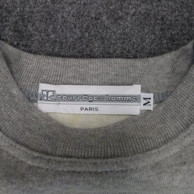 May include: Close-up of a gray sweatshirt collar with a white tag. The tag reads "courreges homme PARIS" with a logo and the size "M". The sweatshirt is a solid gray color.