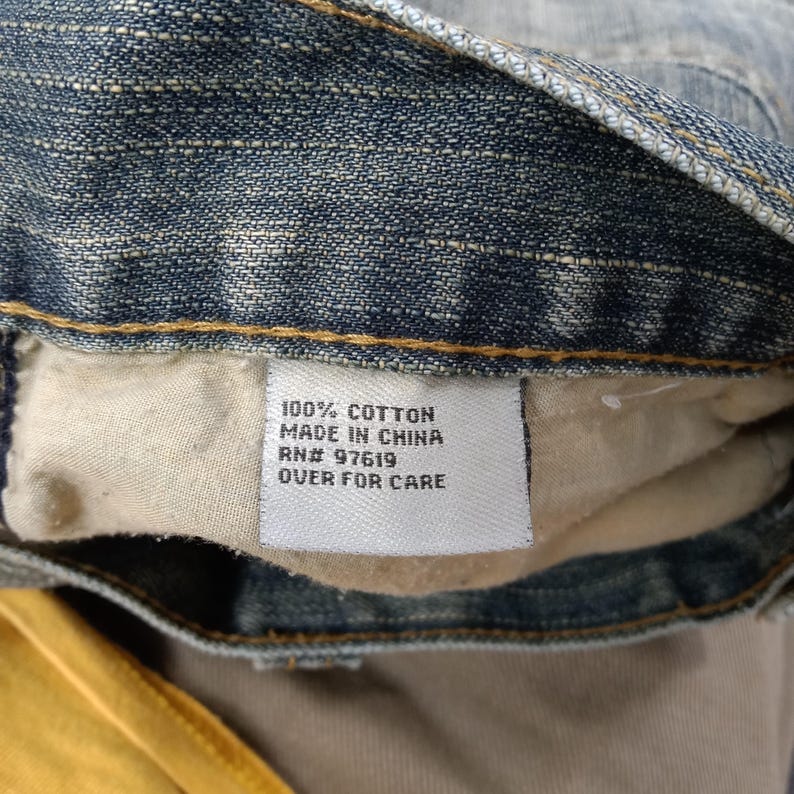 May include: Close-up of a denim garment with a white tag. The tag reads "100% COTTON MADE IN CHINA RN# 97619 OVER FOR CARE". The denim is a light blue wash.