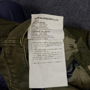 May include: A white tag with black text detailing the care instructions for a garment. The tag reads "COMME des GAR&Ccedil;ONS Co., Ltd." and "MADE IN JAPAN". The tag also includes the fabric content and washing instructions in both English and Japanese.