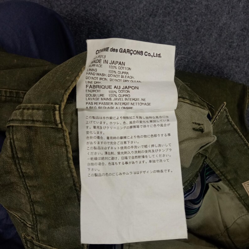 May include: A white tag with black text detailing the care instructions for a garment. The tag reads "COMME des GAR&Ccedil;ONS Co., Ltd." and "MADE IN JAPAN". The tag also includes the fabric content and washing instructions in both English and Japanese.