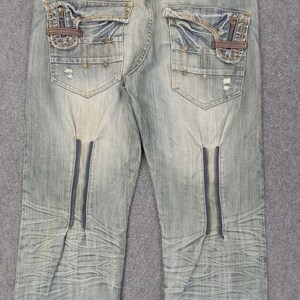 May include: Light blue denim jeans with distressed details, featuring decorative zippers on the front and pockets with zipper and leather accents. The jeans have a faded wash and a classic five-pocket design.